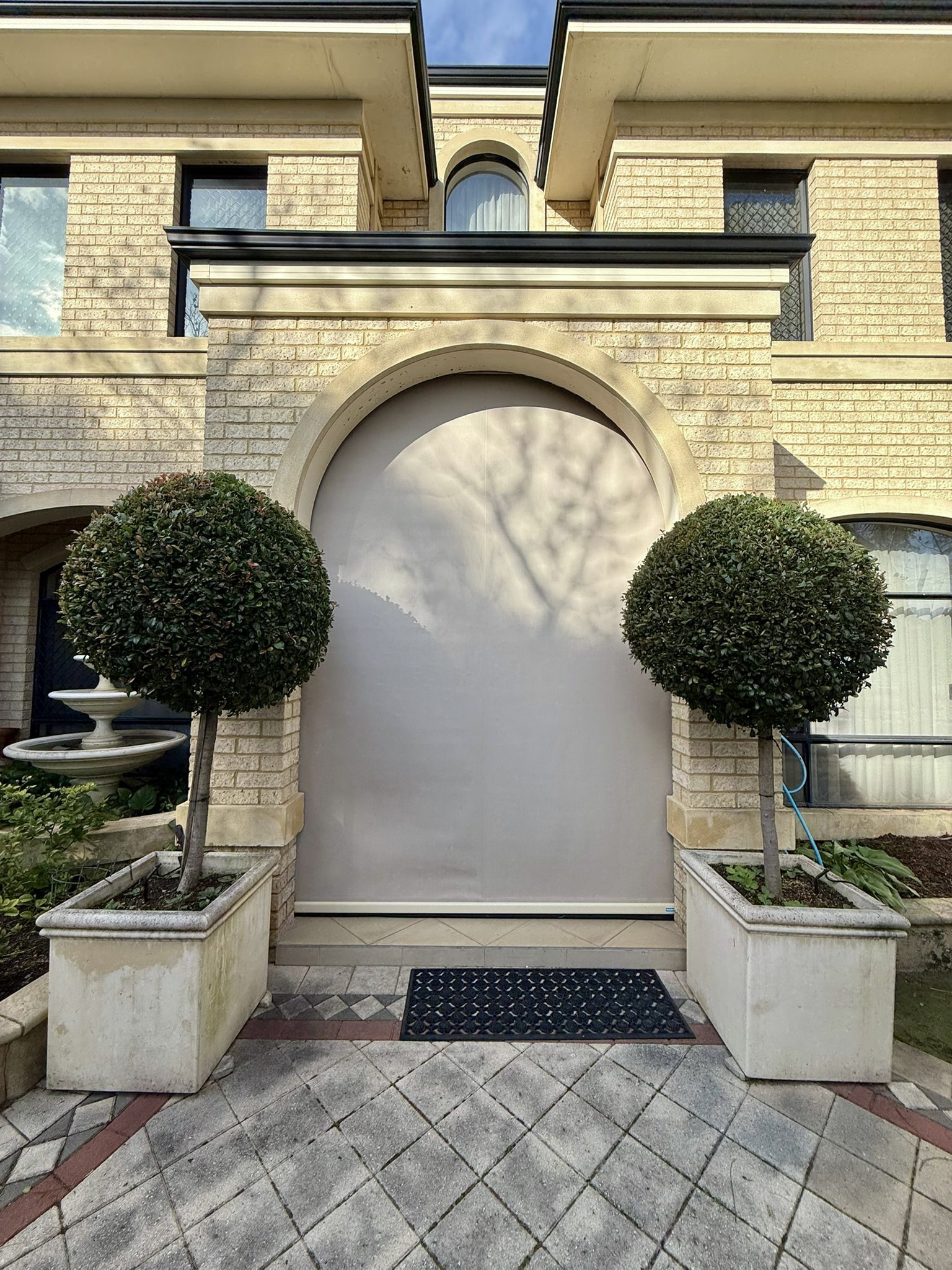 Neutral outdoor blind lowered over an arched entry of a traditional Perth home with topiary and stone detailing