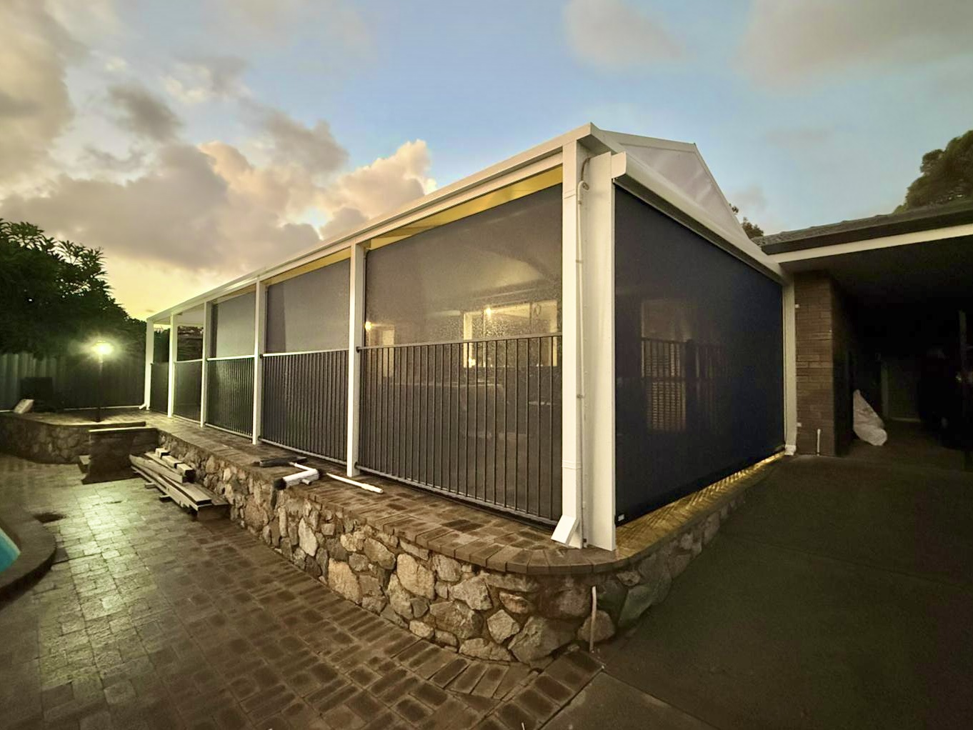 Charcoal mesh outdoor blinds fully lowered across a large covered patio in Perth at dusk, with a pool area and stone retaining wall