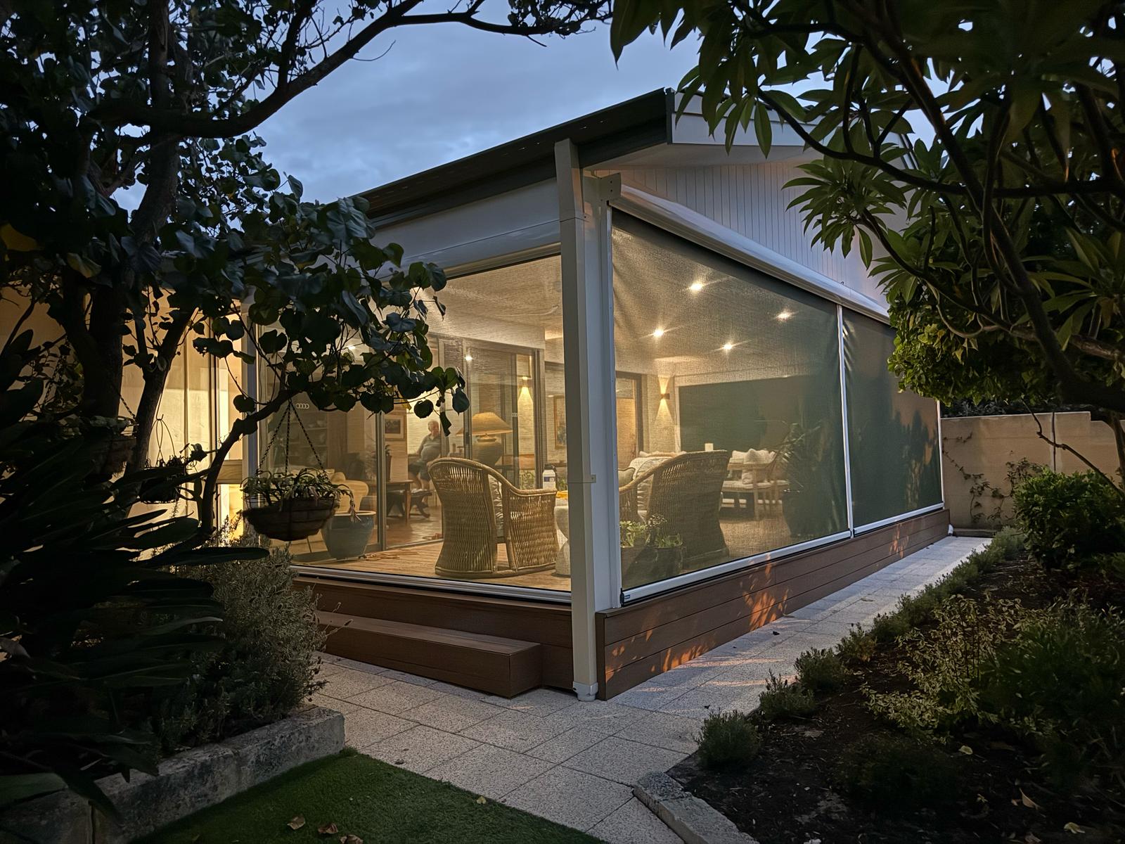 Mesh outdoor blinds fully enclosed around a Perth home's outdoor entertaining area at dusk, with warm interior lighting visible through the fabric