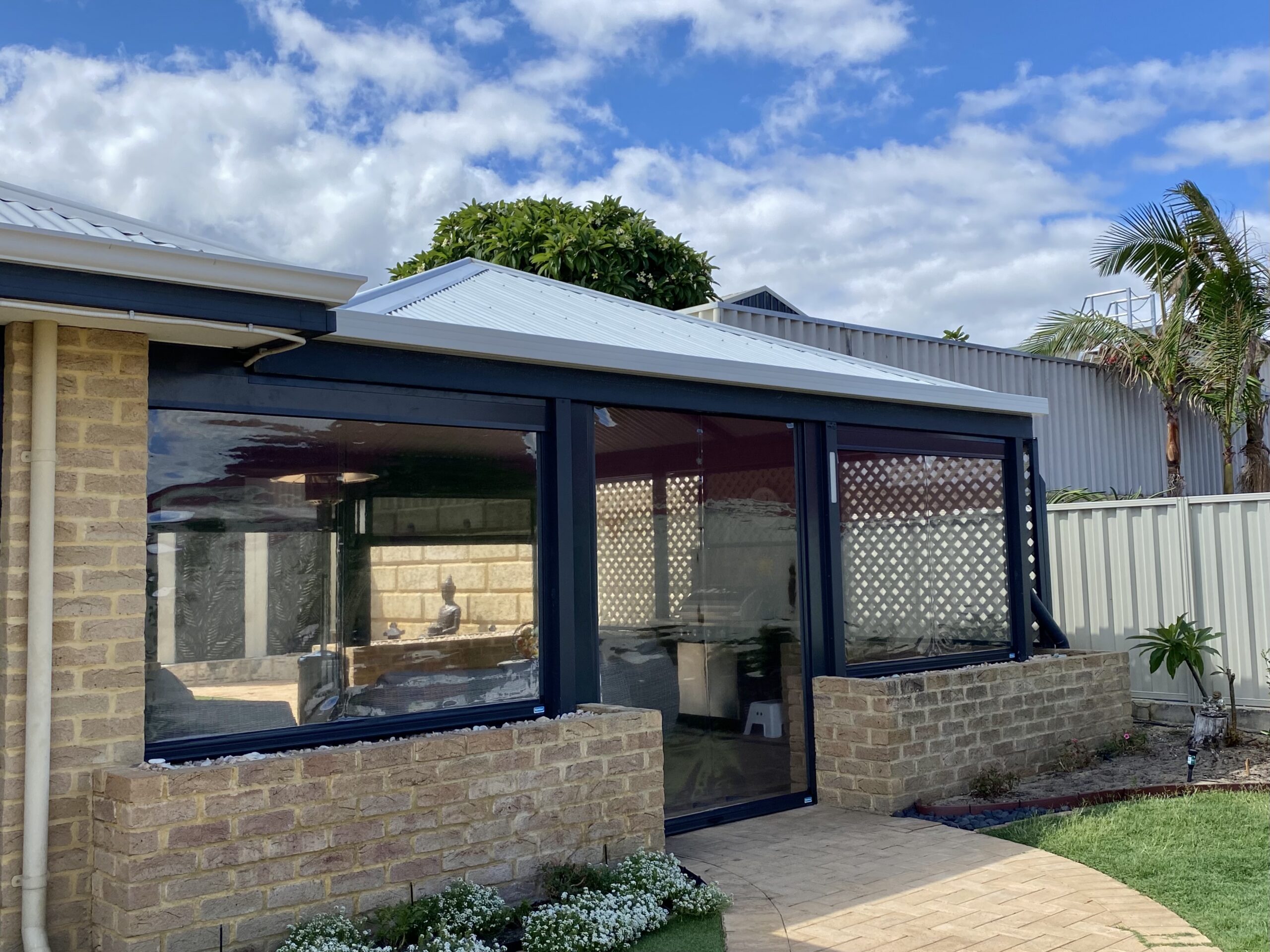 Clear PVC outdoor blinds fully lowered on the exterior of a brick Perth home with a dark grey track system and blue sky